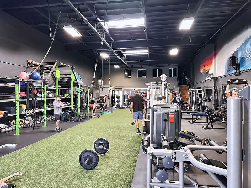 Long view of the gym with a turf lane, weight plates, strength machines and overhead lighting.